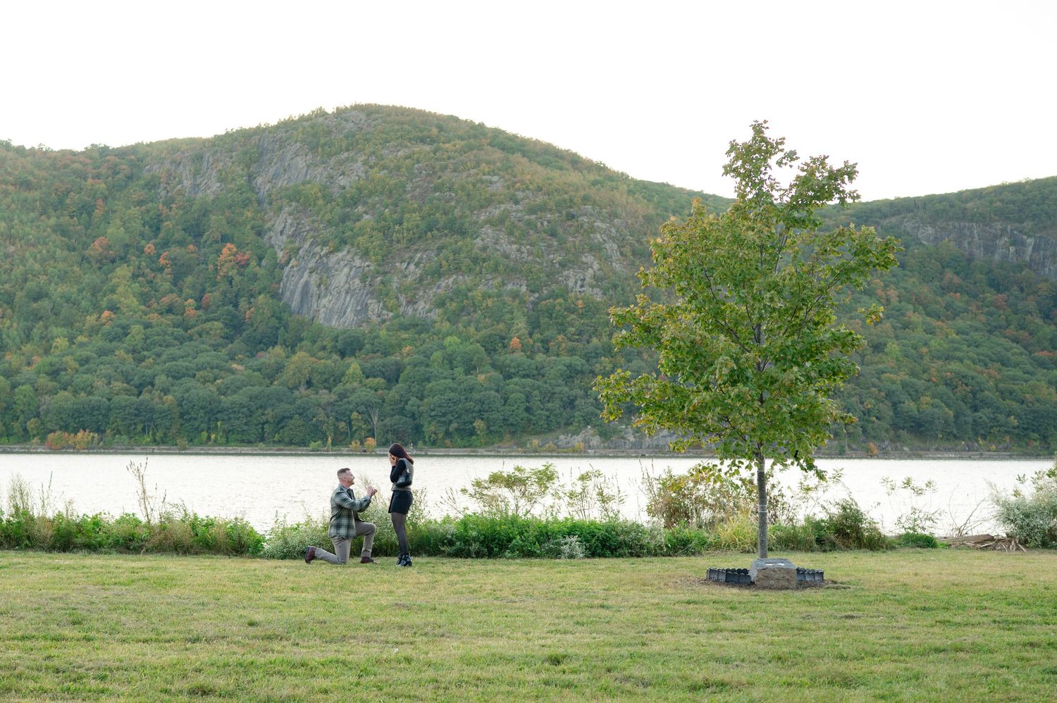 Person kneeling in a marriage proposal on a grassy lawn beside the Hudson River, with a  mountain in the background in Cold Spring, NY.
