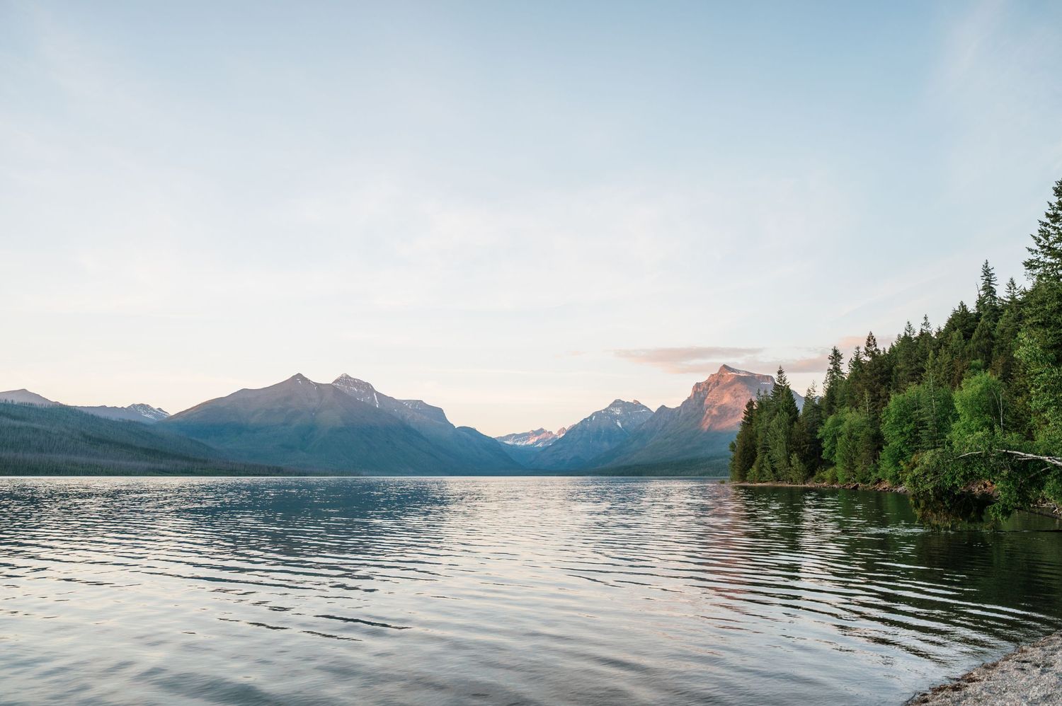 Serene mountain lake surrounded by evergreen trees with snow-capped peaks reflecting in calm water at sunset.