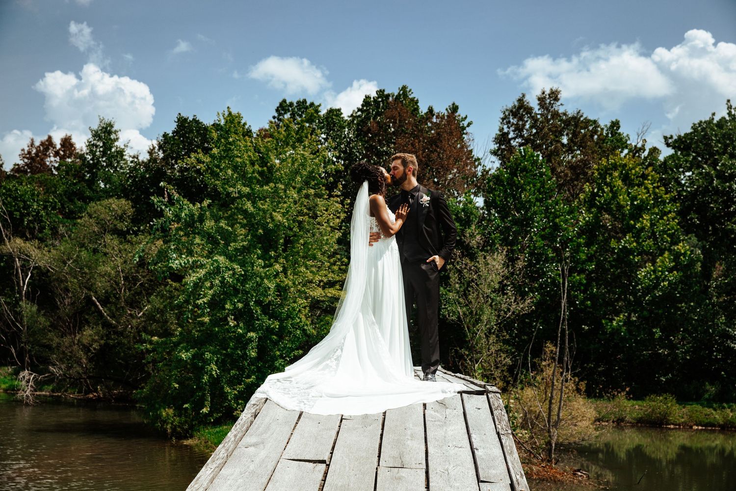 Bride and groom kissing on a wooden dock surrounded by lush trees and a serene lake on their wedding day.