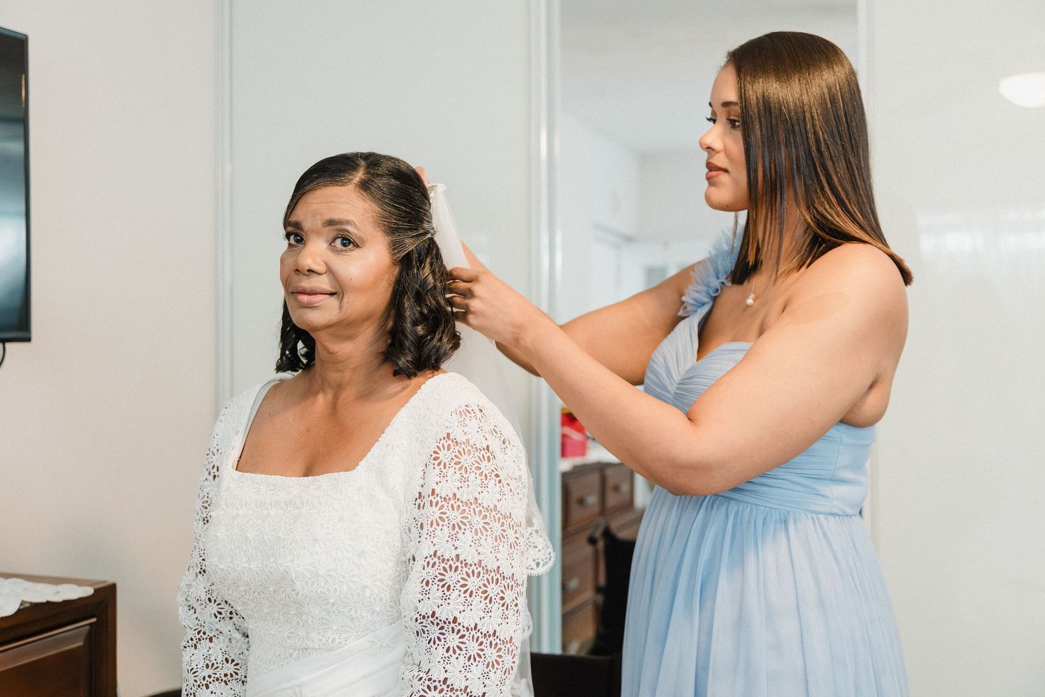 Una mujer con vestido azul arregla el cabello de la novia, vestida de blanco, durante sus preparativos.