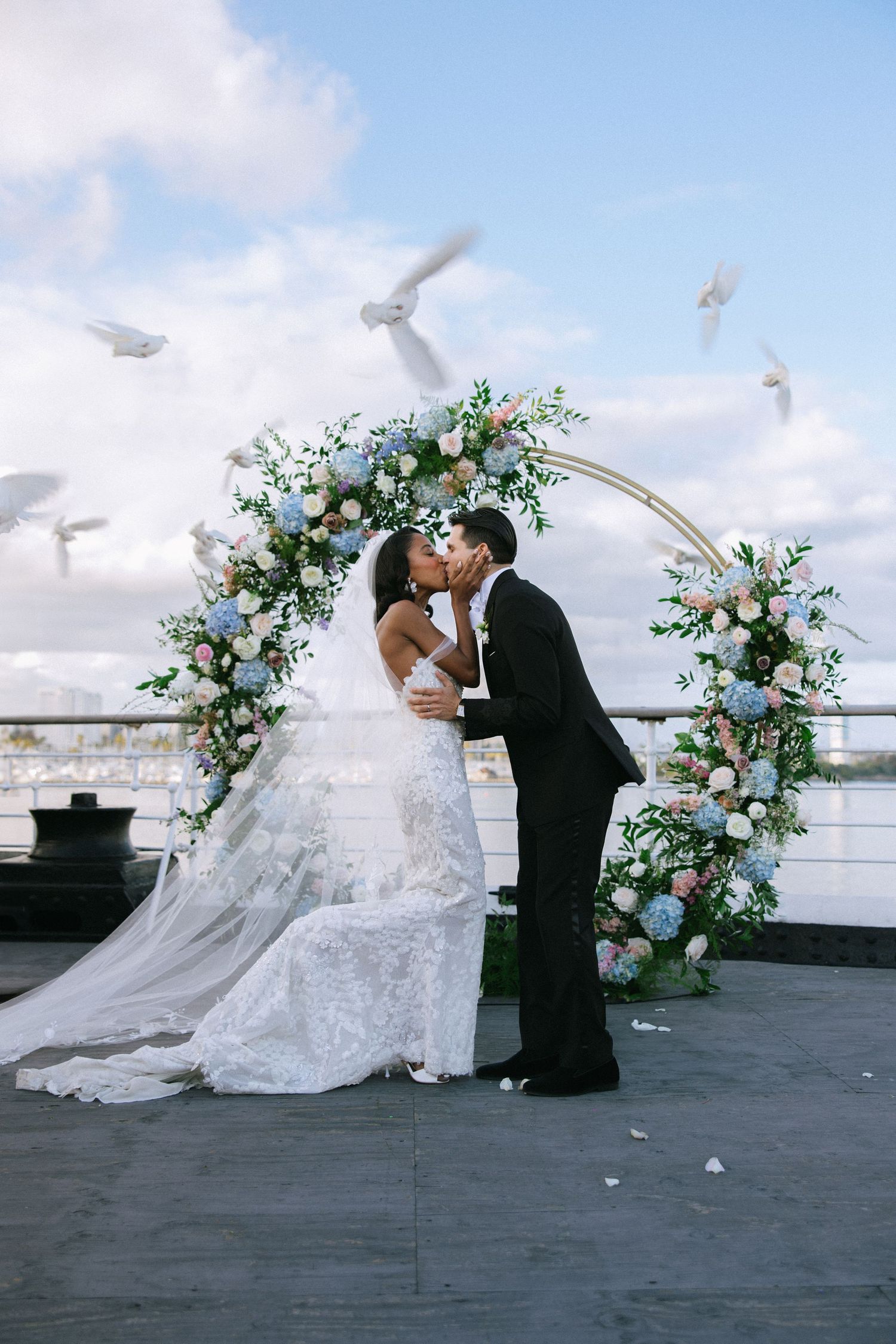 Bride and groom kissing under floral arch with white doves flying at waterfront ceremony.