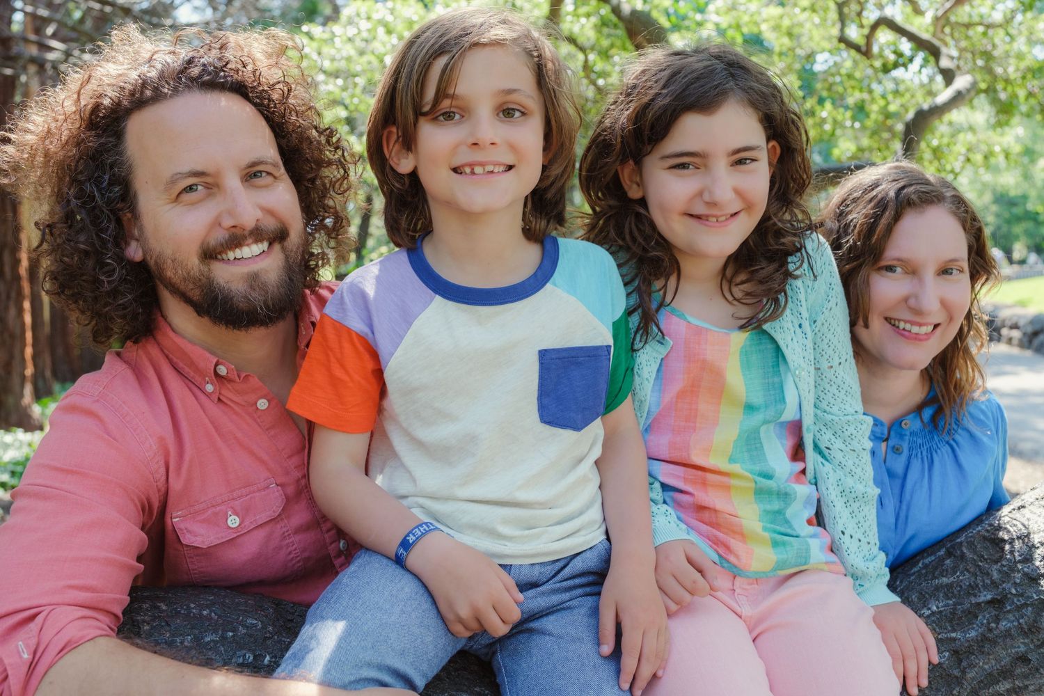 A family shares a joyful moment together while sitting on a large tree trunk in a sunlit outdoor setting.
