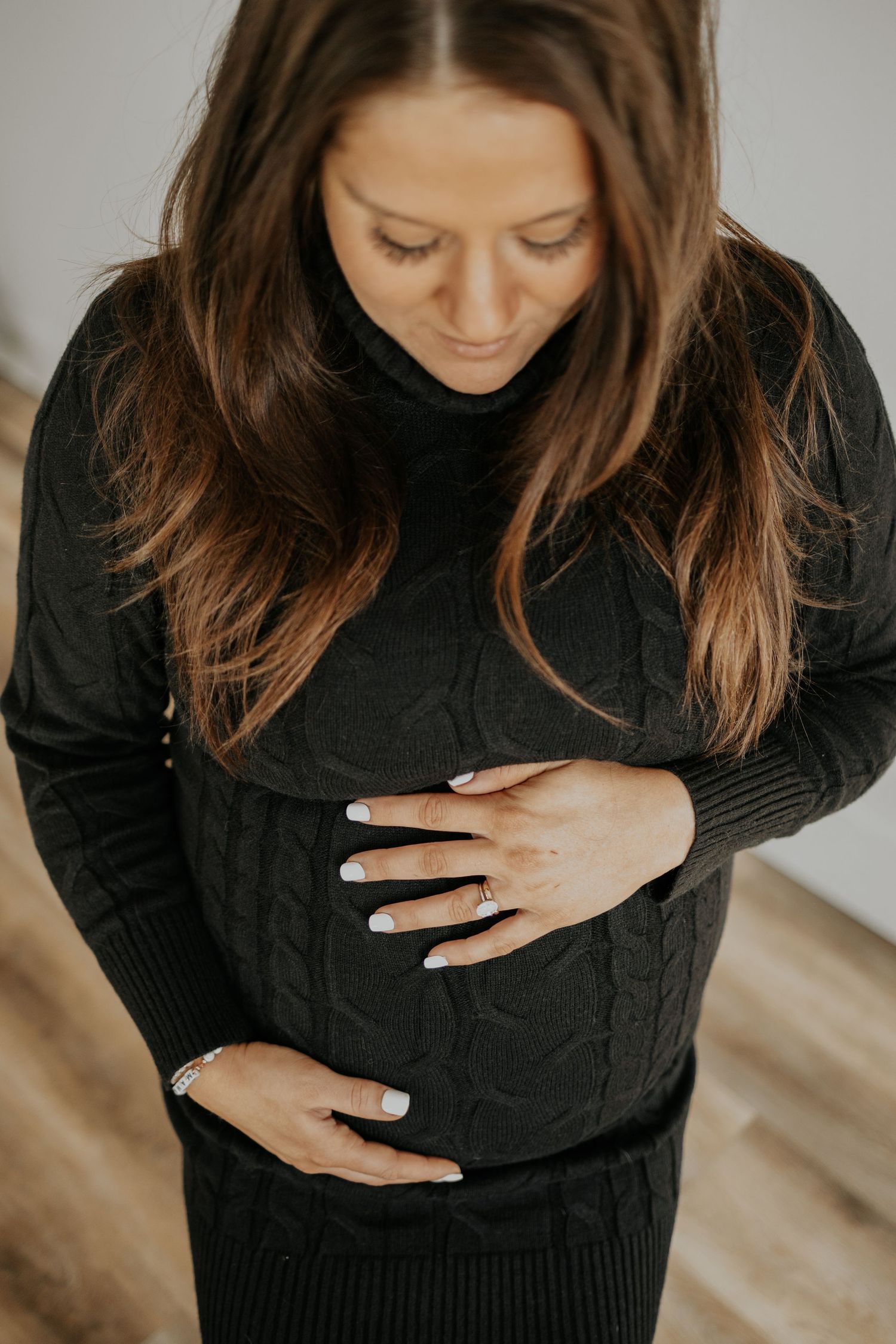 A person in a black sweater gently places their hands on their stomach while looking down, suggesting pregnancy.