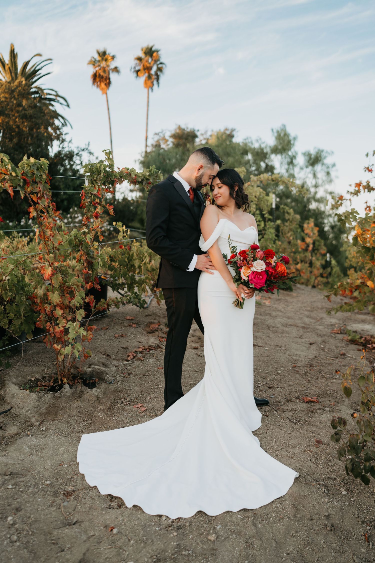 Wedding couple embraces on dirt path with palm trees and red floral bouquet against sunset sky in tropical garden setting.