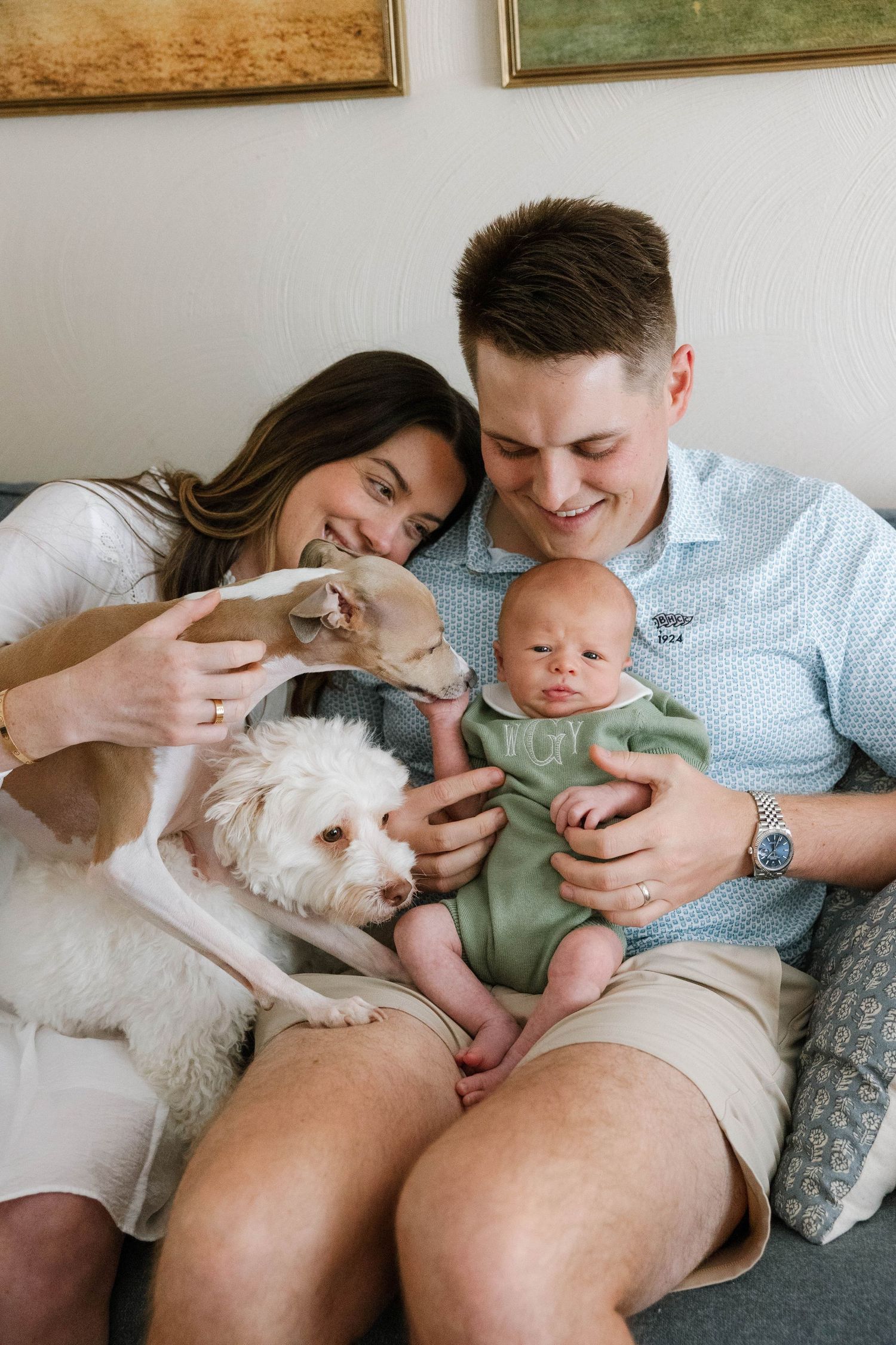 A couple snuggles with a small infant and white fluffy dog on a cozy gray couch.