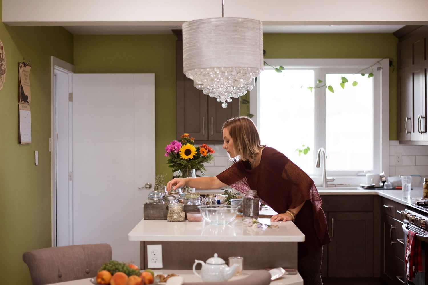 Person reaching across kitchen counter with modern chandelier and green walls.