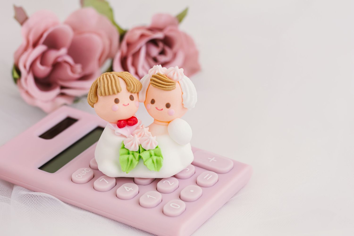 Miniature wedding cake topper figures sit on a pink calculator with pink roses in the background.