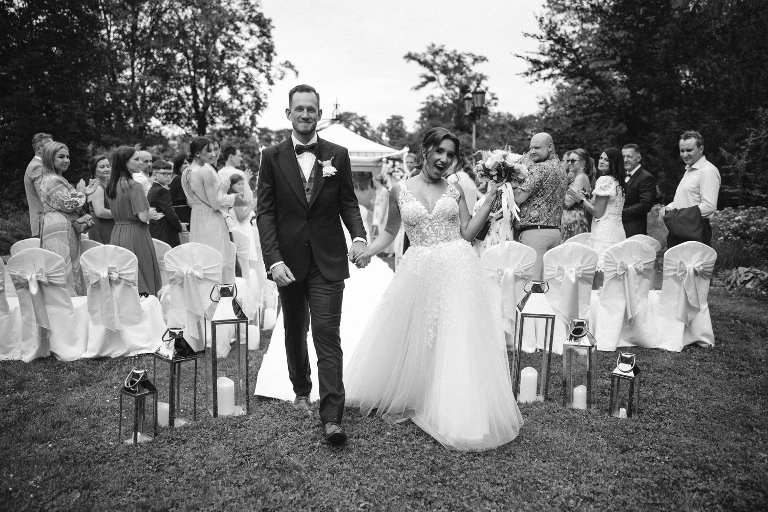 Black and white photo of newlyweds walking down the aisle lined with lanterns at an outdoor ceremony.