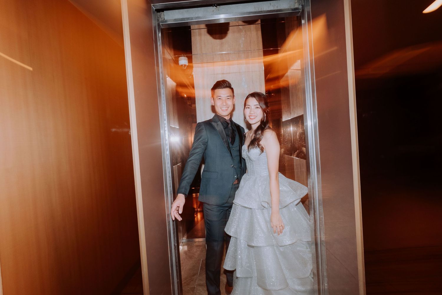 A couple in formal attire pose together inside an elevator, with warm lighting and wood paneled walls.
