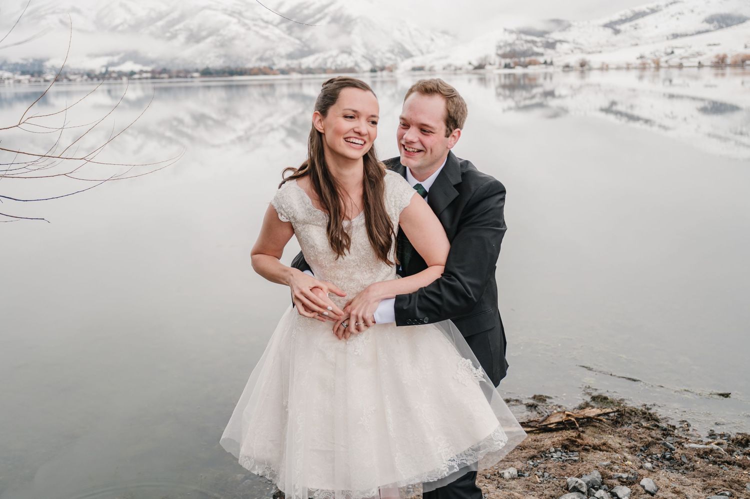 A couple in wedding attire embrace and laugh together by a snow-covered mountain lake on their winter wedding day.