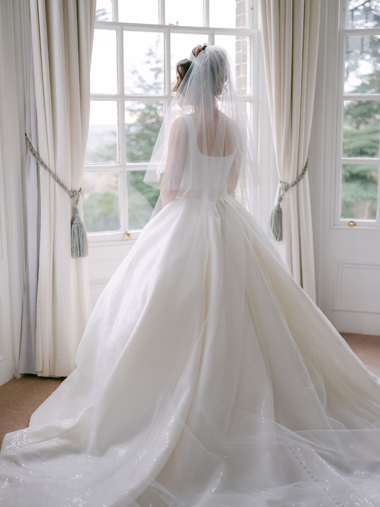 Elegant white wedding gown with flowing train photographed from behind near a bright window.