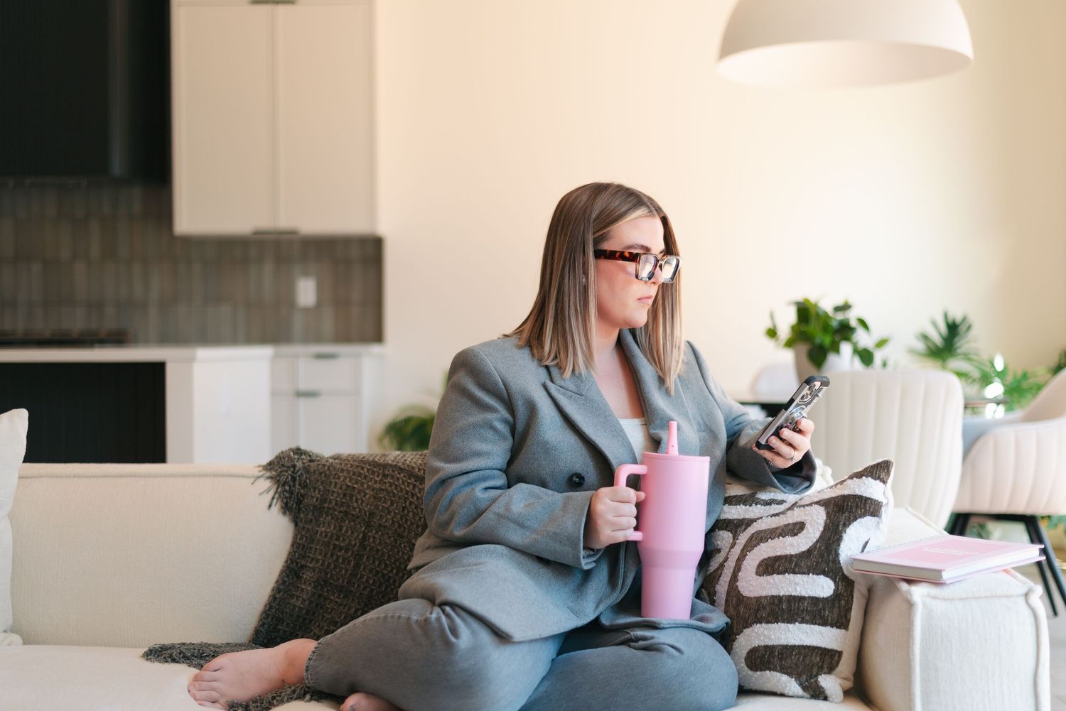 Professional in a gray blazer sits on couch with pink smoothie while checking smartphone in modern home office.