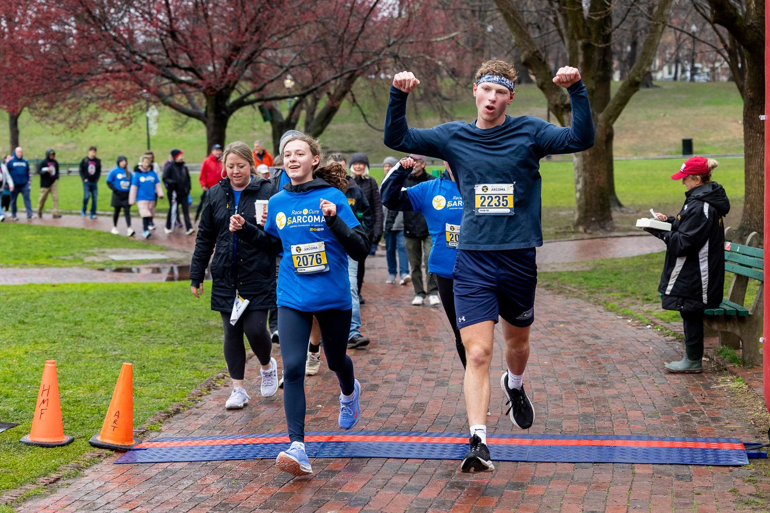 Athletes running across blue timing mat on path during outdoor race with orange cones and spectators in background.