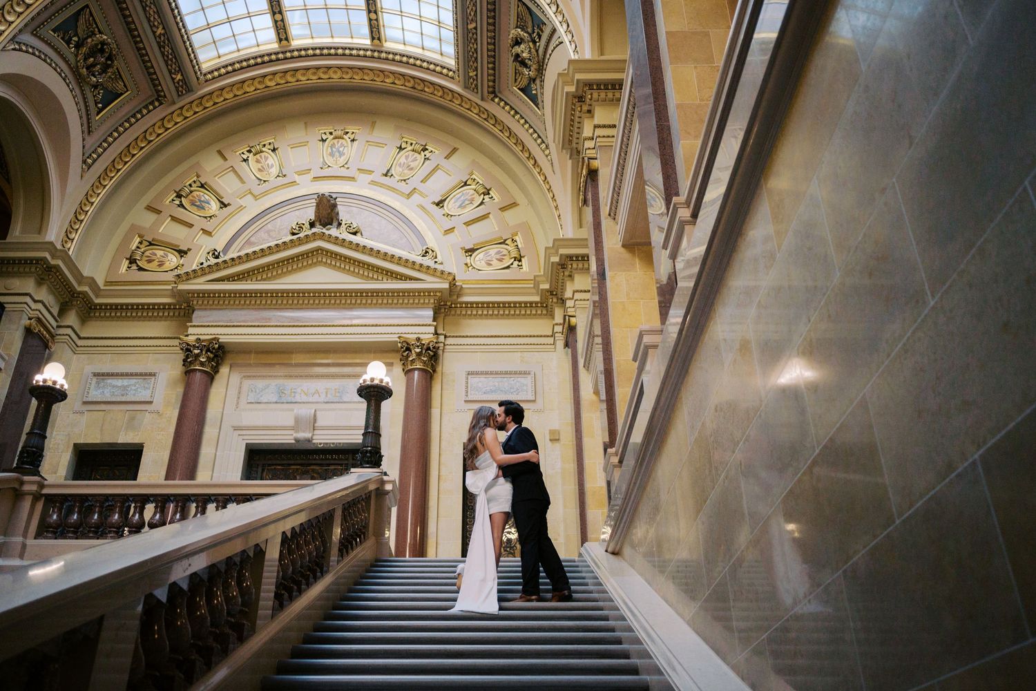 A couple shares a romantic moment on an elegant marble staircase inside a grand historic building with ornate architecture.