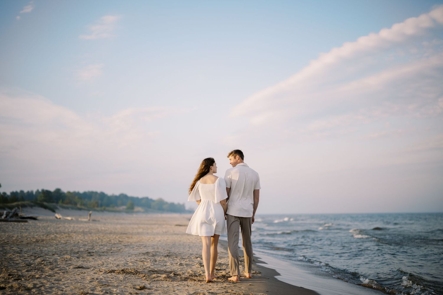 A couple walking along the shoreline on a sandy beach at sunset with soft pink skies and rolling waves.