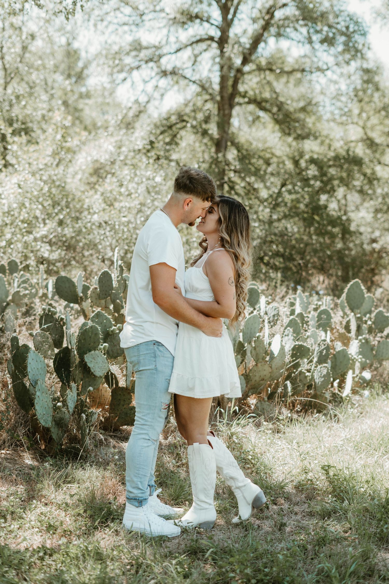 A romantic couple embraces in a rustic setting with blooming cactus and trees in the background.