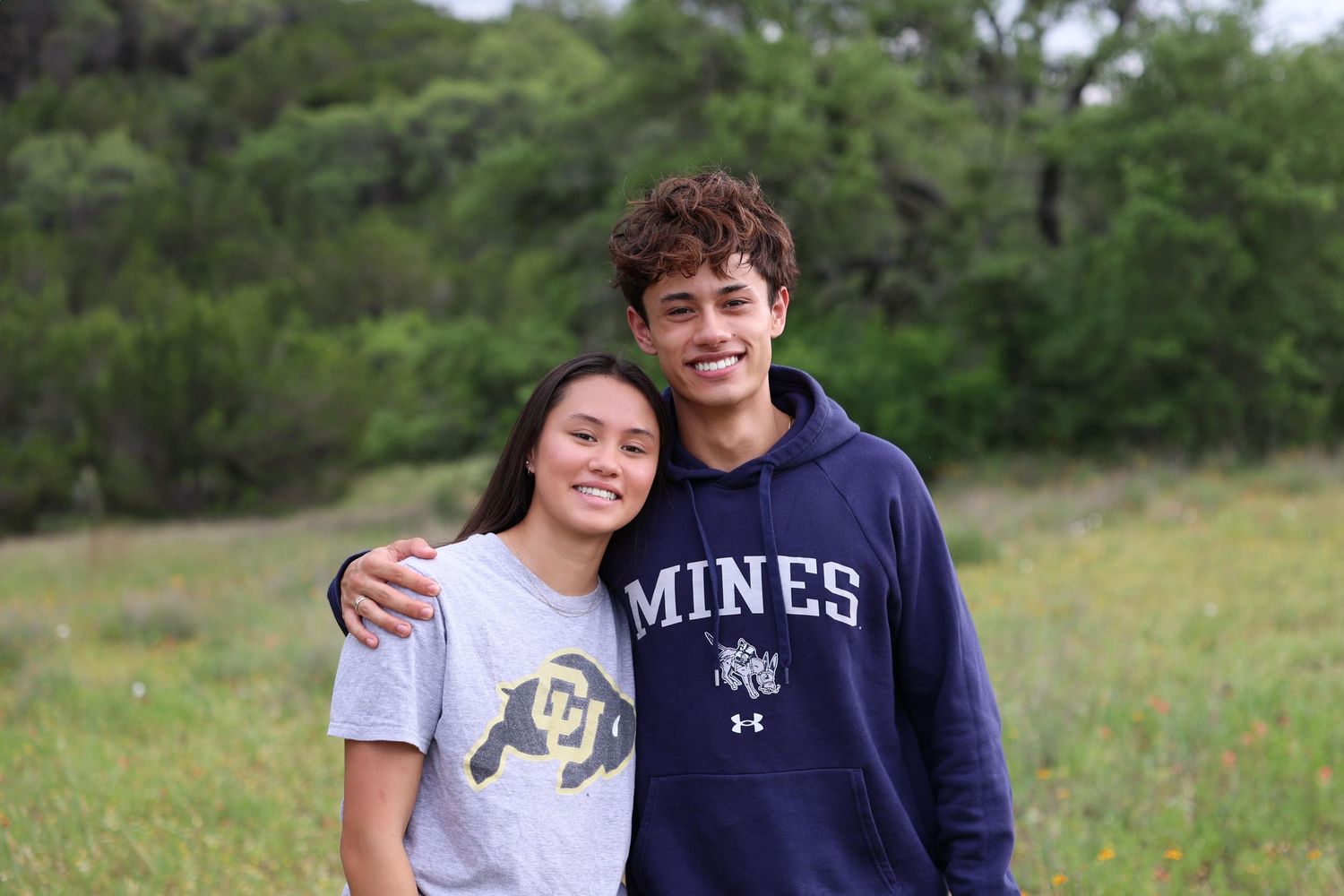 Two students stand together in a grassy field with trees in background, one wearing a Mines college sweatshirt.