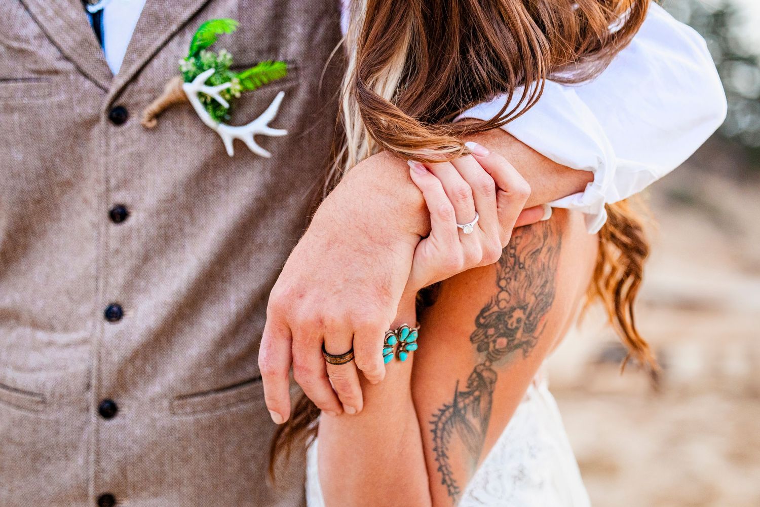 Close up of holding hands showing tattoo artwork and turquoise ring against brown suit jacket with floral boutonniere.