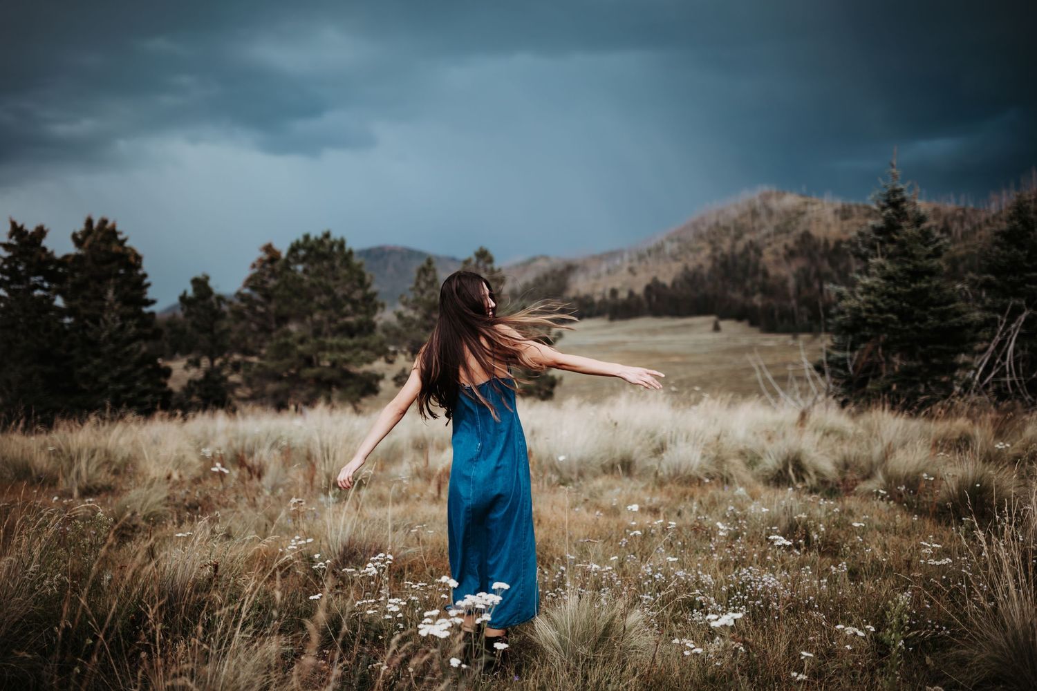 Woman in blue dress stands joyfully in mountain meadow under stormy skies.