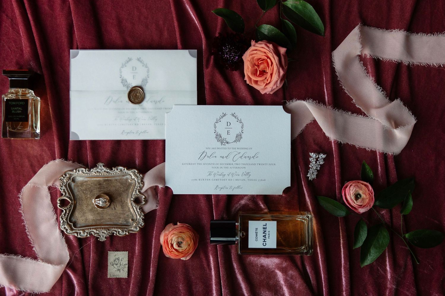 Wedding invitation suite with wax seal, roses, and perfume bottles arranged on burgundy fabric.