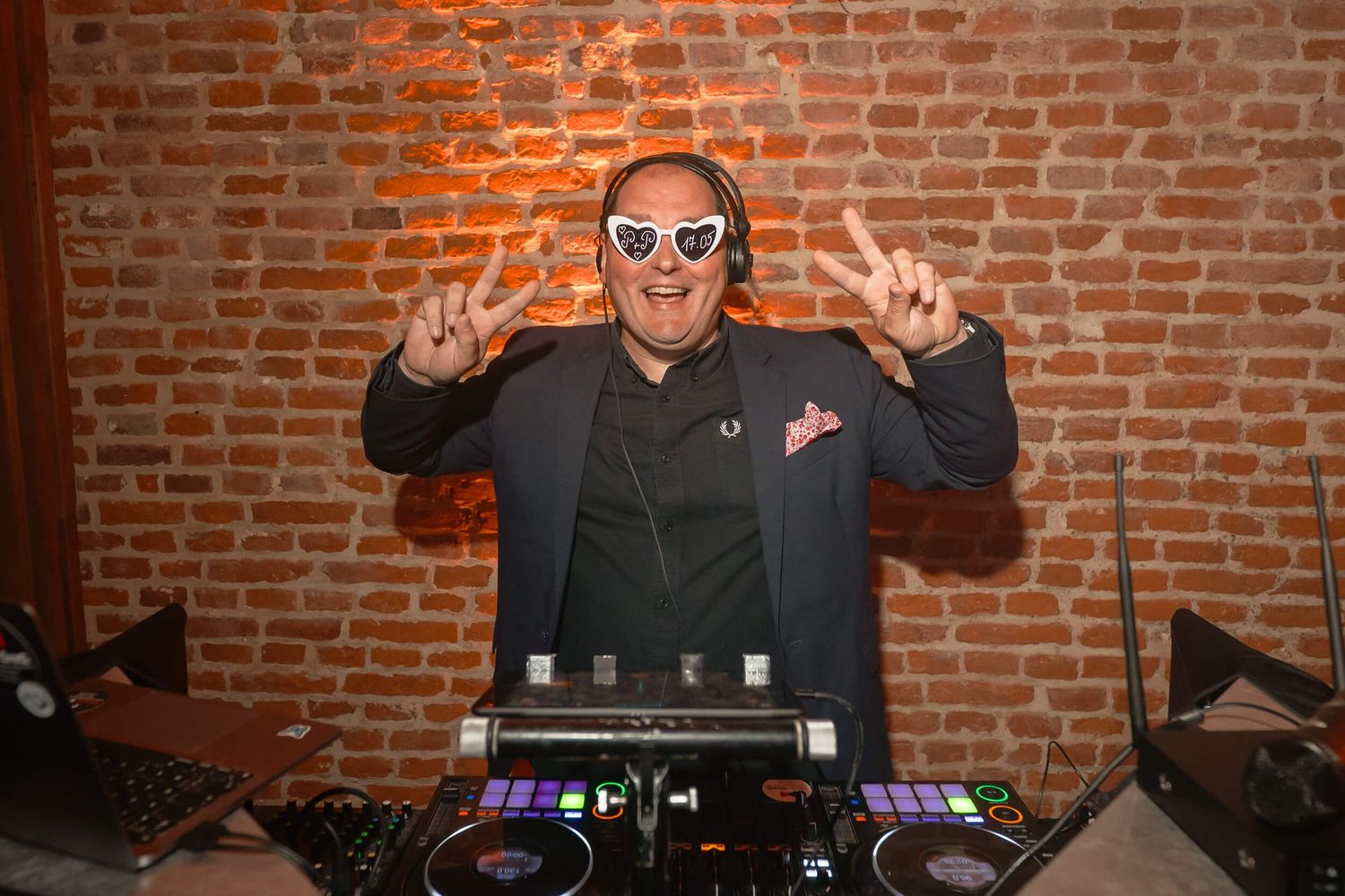 DJ performing at a venue with brick wall backdrop showing peace sign gesture while standing at mixing equipment.