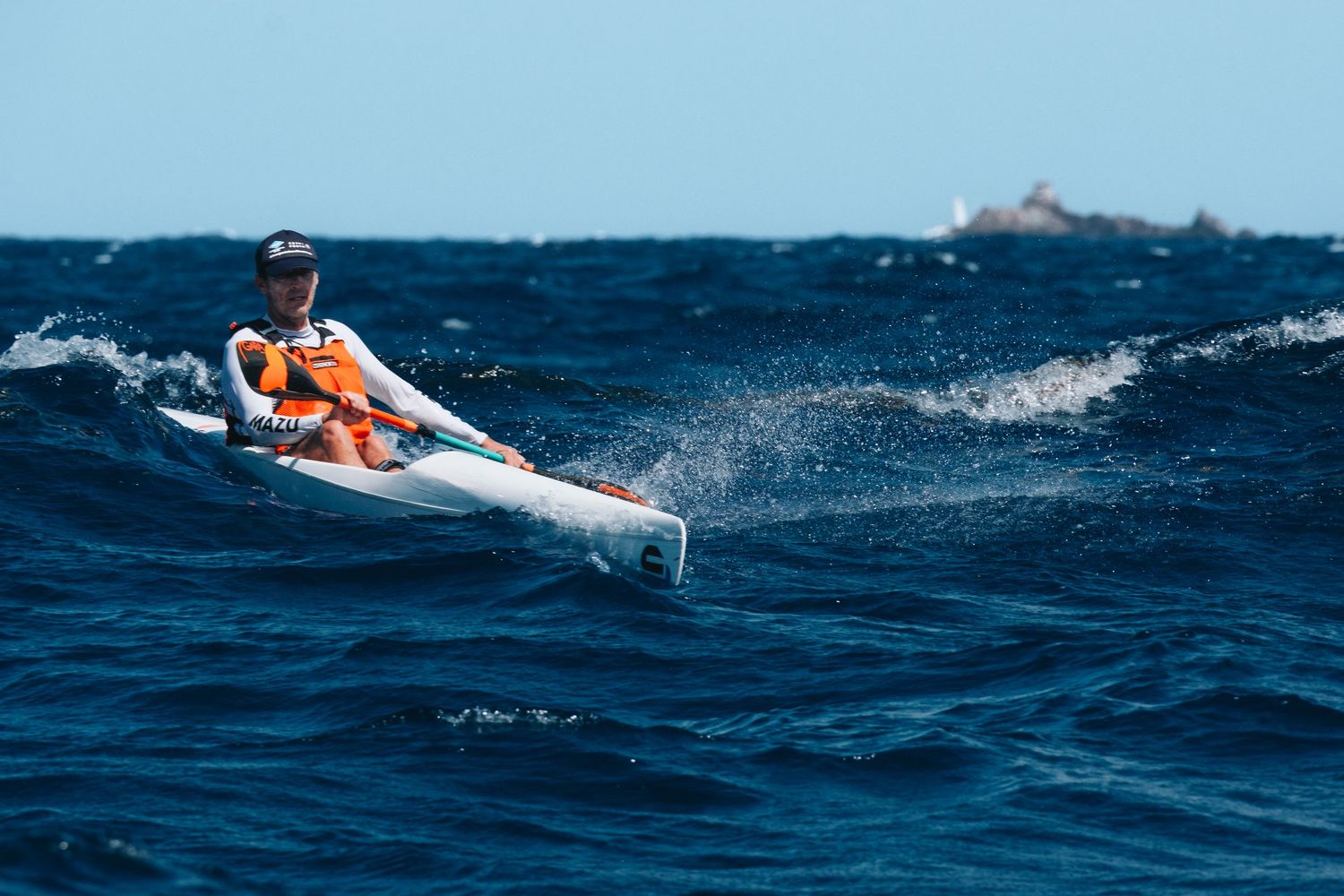 A kayaker navigates through choppy blue ocean waters with a distant island visible on the horizon.