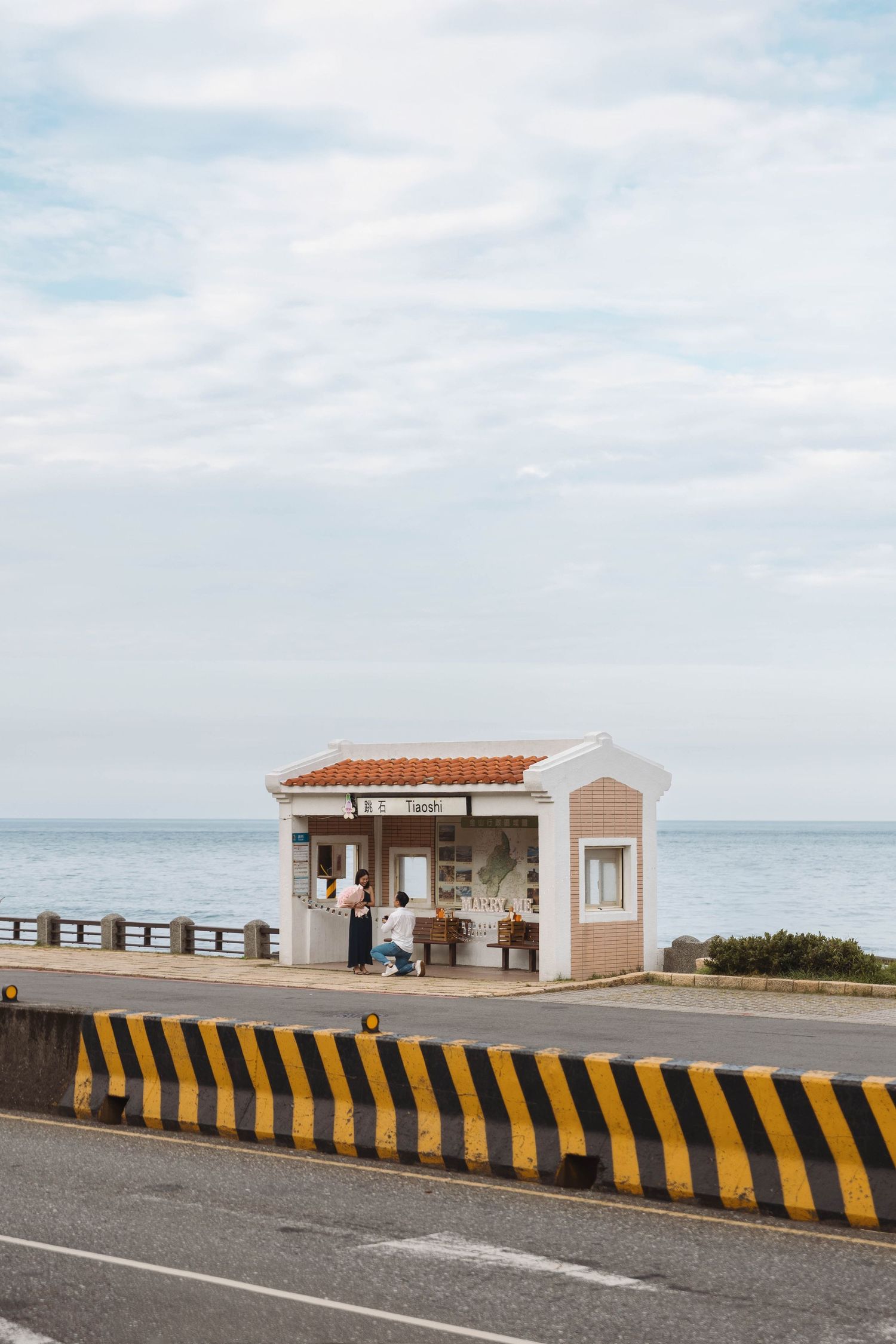 Small white seaside bus stop kiosk along coastal highway with ocean view under cloudy sky.