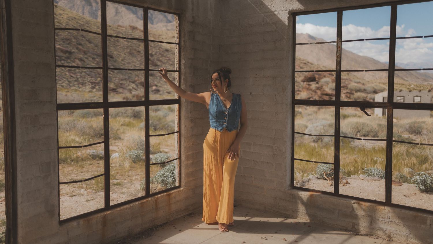 Person in blue top and yellow pants stands by large windows in a desert building interior.
