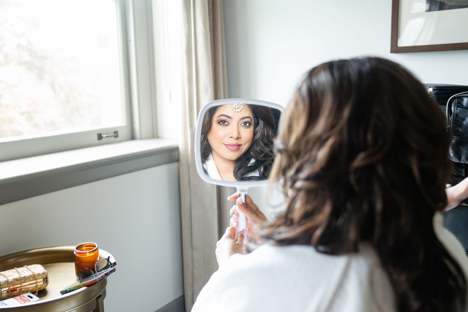 Woman holding a small mirror reflecting her face while getting ready near a bright window.