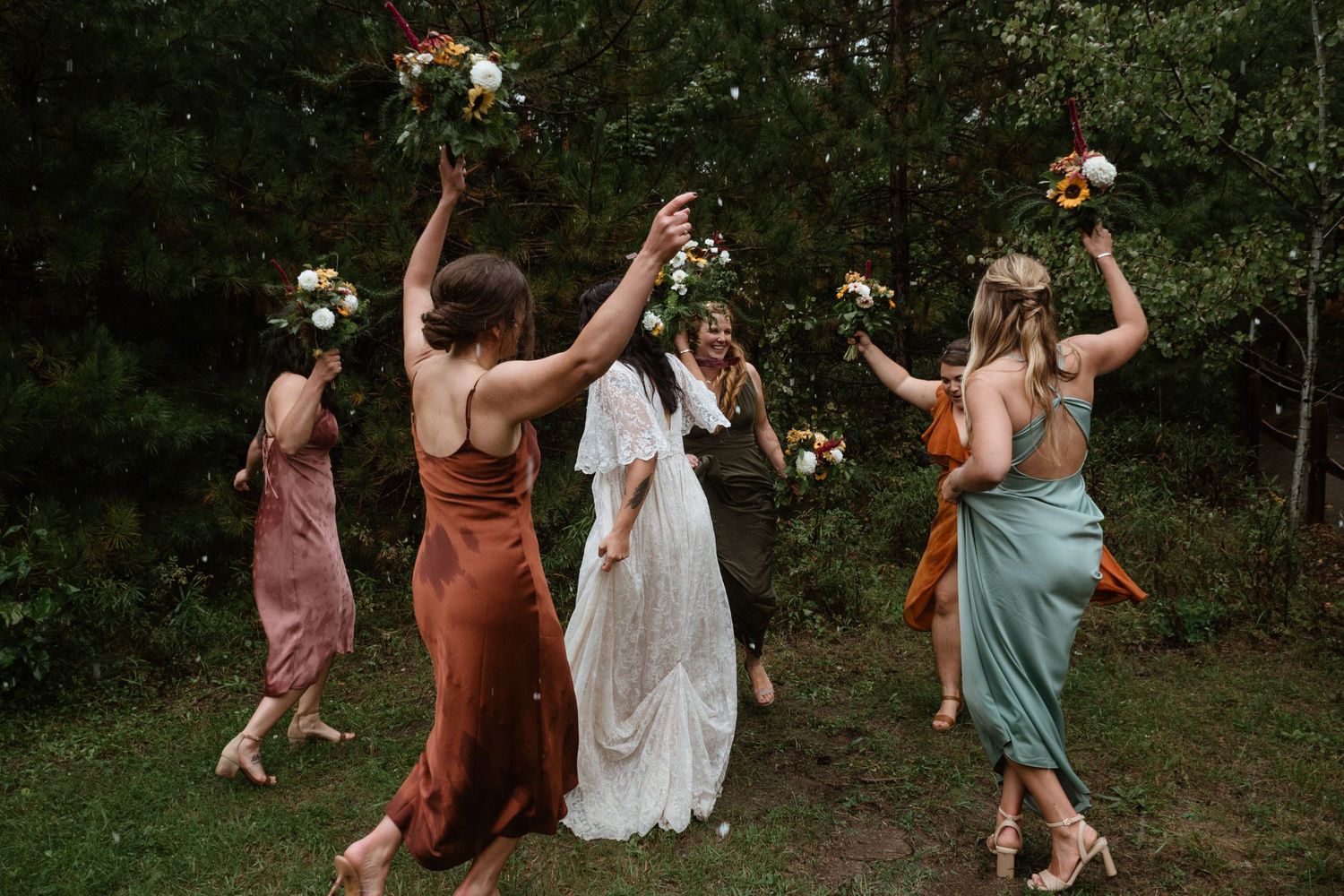 Bridesmaids in colorful silk dresses dance joyfully while holding bouquets in a garden setting.
