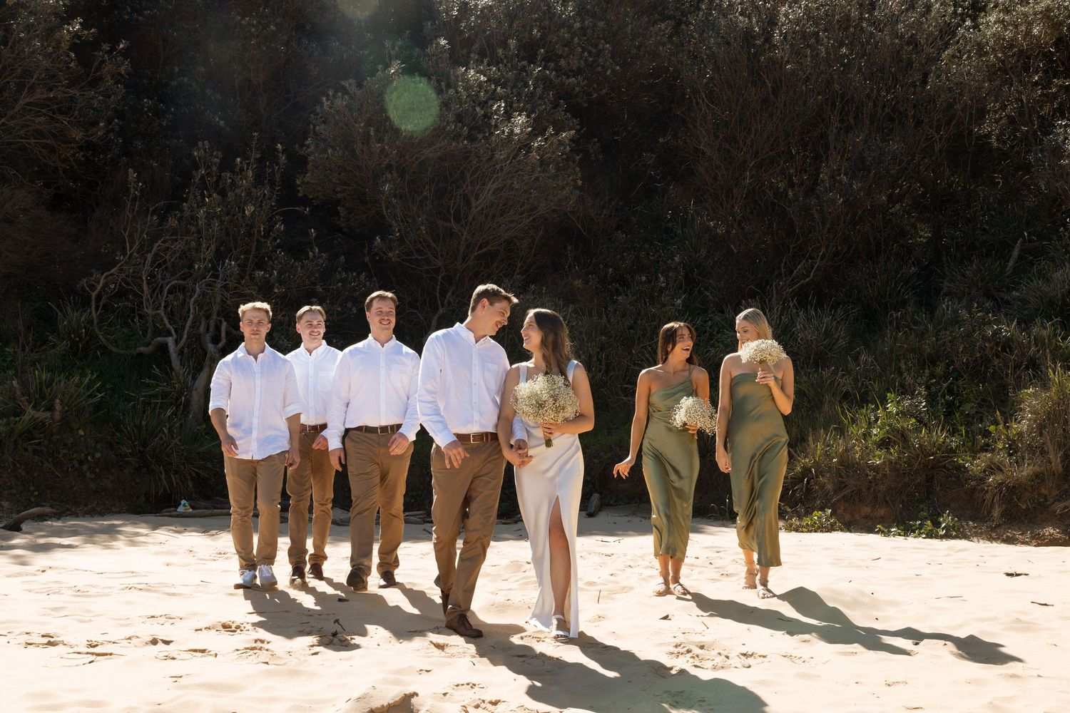 Wedding party in coordinated outfits poses on beach with natural sunlight.