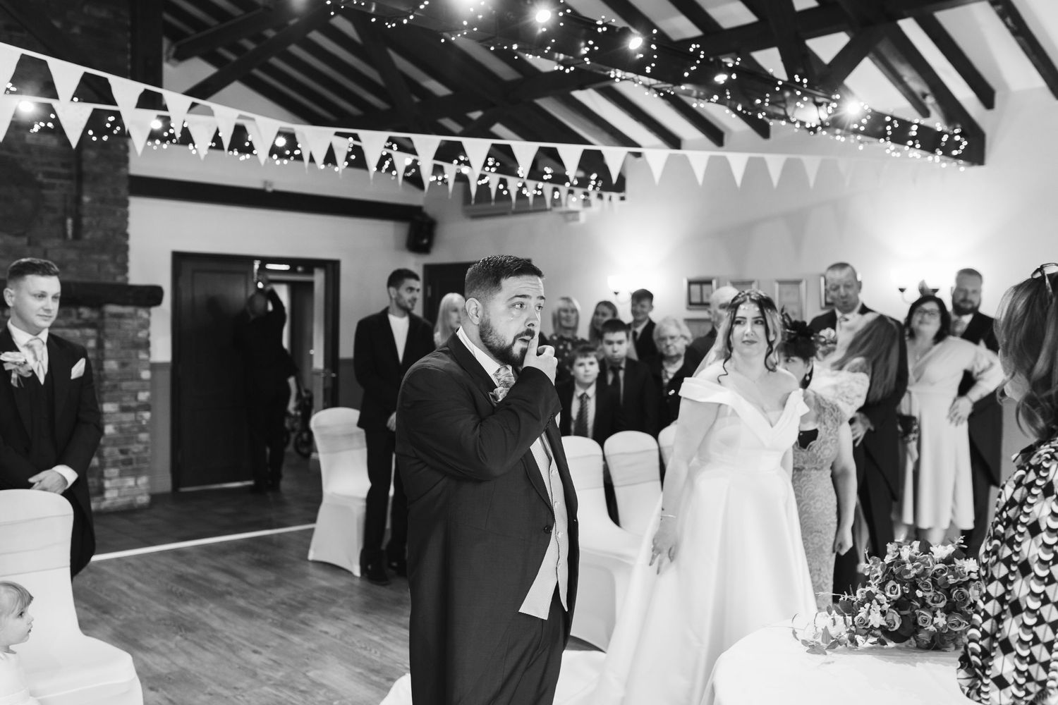 Black and white photo of a wedding reception with a groom speaking into a microphone while guests watch from the sides.