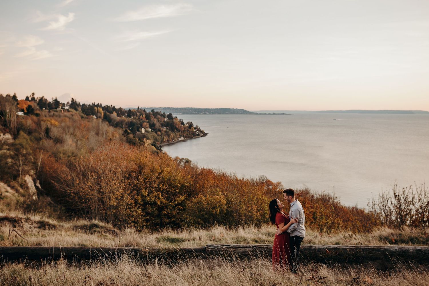 Couple embraces on cliffside overlooking water during sunset with autumn foliage along Pacific Northwest coastline.