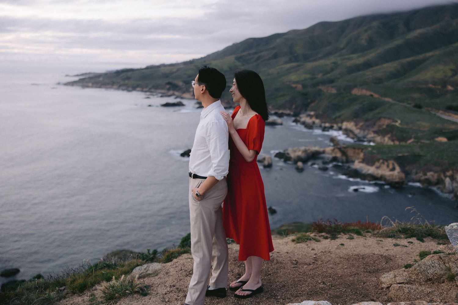 Couple standing on coastal cliff overlooking dramatic Big Sur coastline with rocky shores and mountains in California.