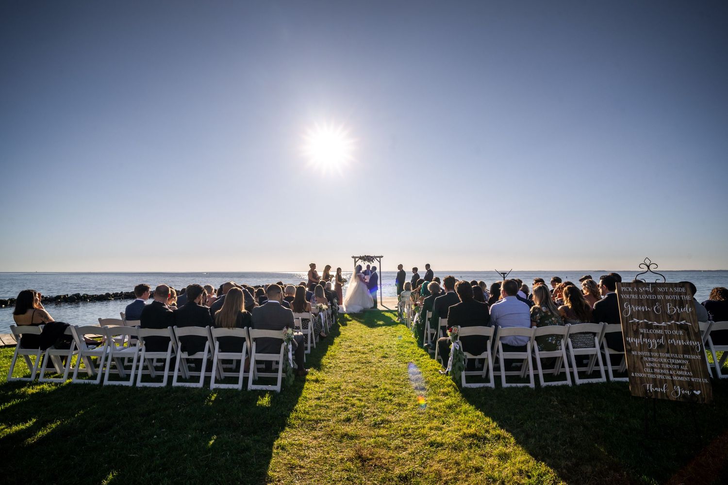 An outdoor wedding ceremony setup with white chairs under bright sunlight by the water.