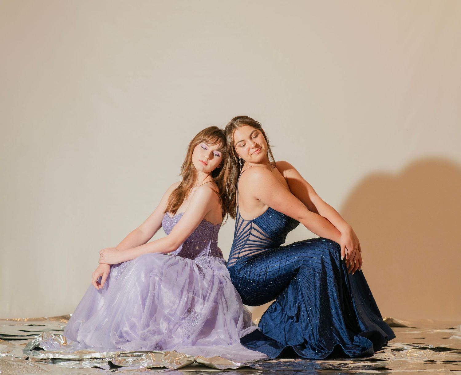 Two young women in elegant prom gowns, lavender and navy blue, sitting back to back on studio floor.