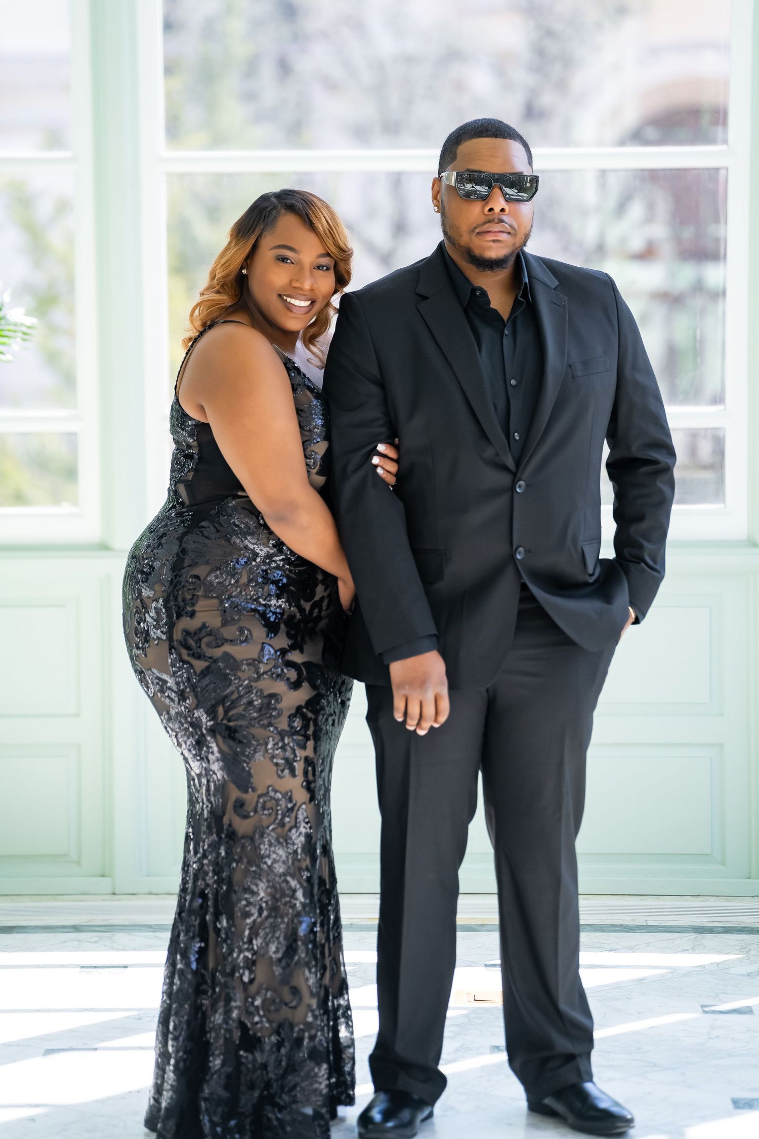 Elegant formal couple photo featuring black evening wear against bright interior backdrop.
