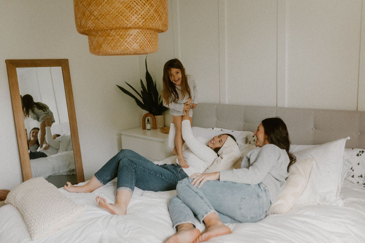 Friends relaxing on comfortable bed with rattan lamp and mirror decor in bright room.