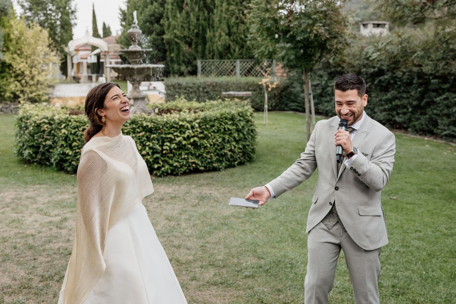 Bride in white gown with cape and groom in gray suit pose on green lawn with tall evergreen trees backdrop.