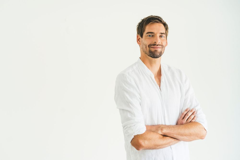 A confident smiling man in a white linen shirt standing with arms crossed against a white background.