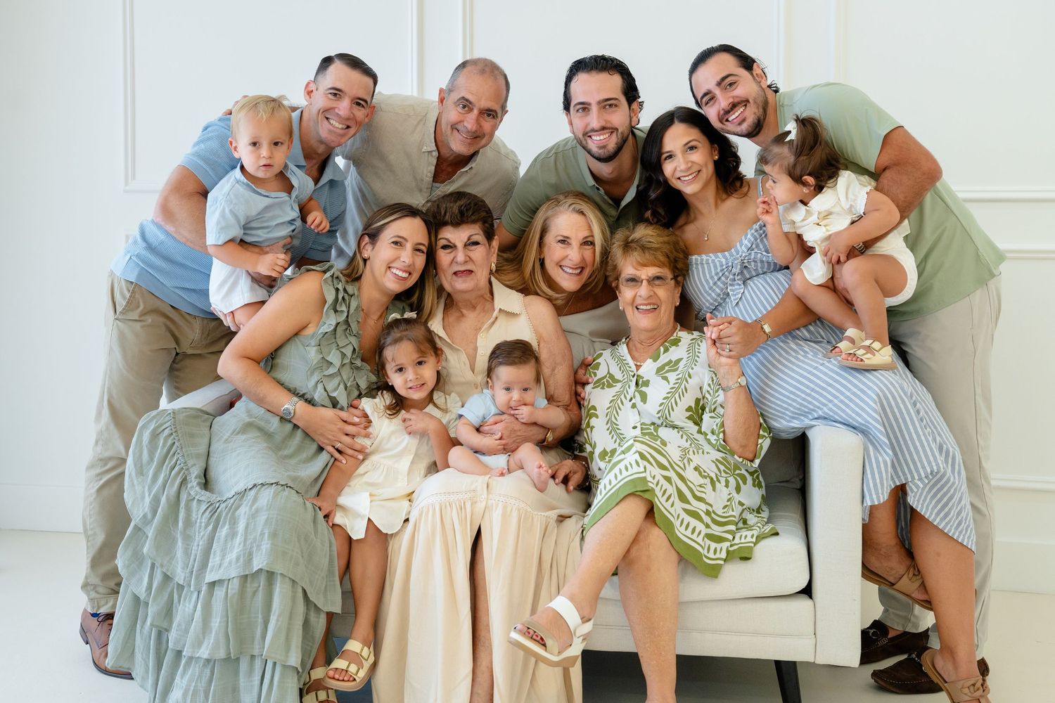 Large multigenerational family portrait with adults and young children smiling together on a white sofa in a bright room.