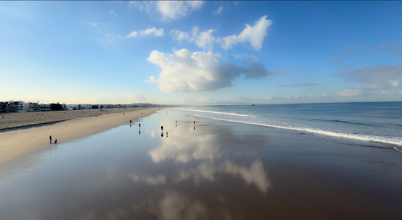 Serene beach with wet sand reflecting clouds and blue sky, gentle waves, and distant figures walking along the shore.