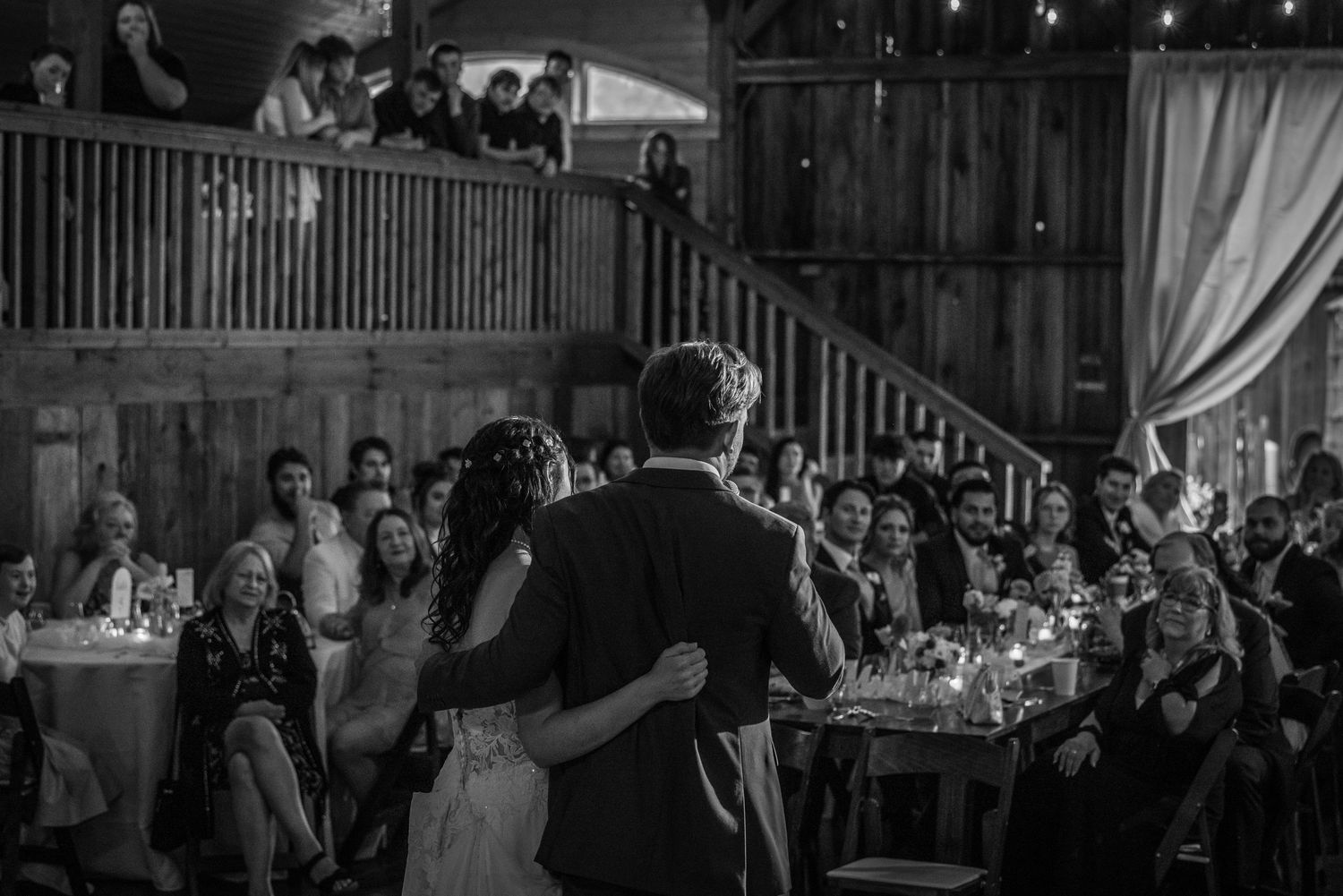 Bride and groom share first dance in rustic barn venue with guests watching from balcony and tables below.