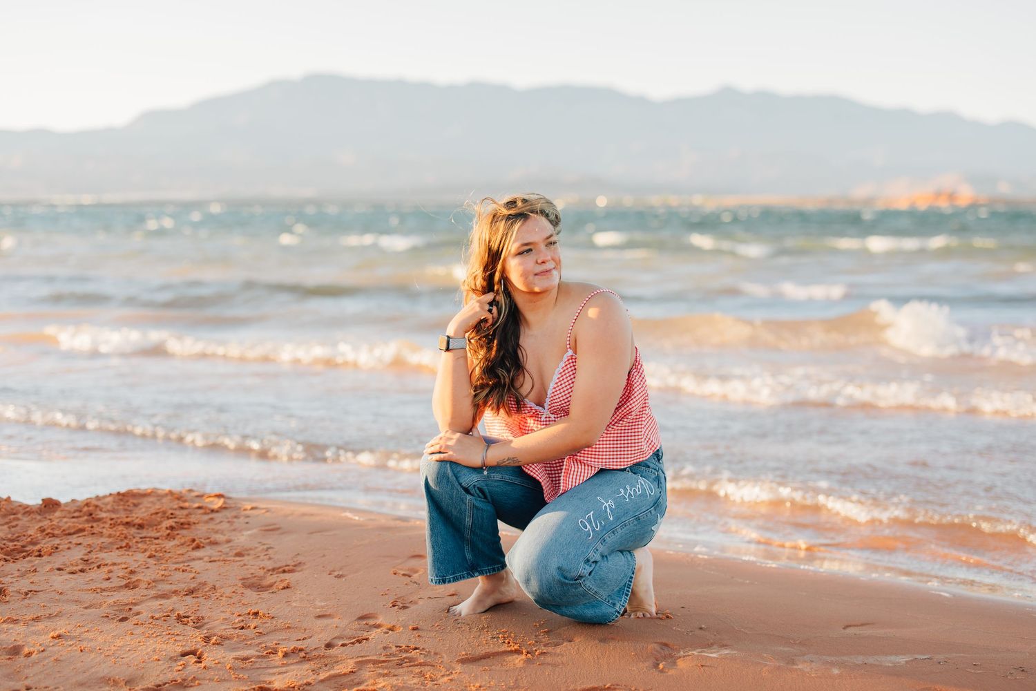 Woman in pink top and jeans posing on a beach with mountains and blue water in the background.