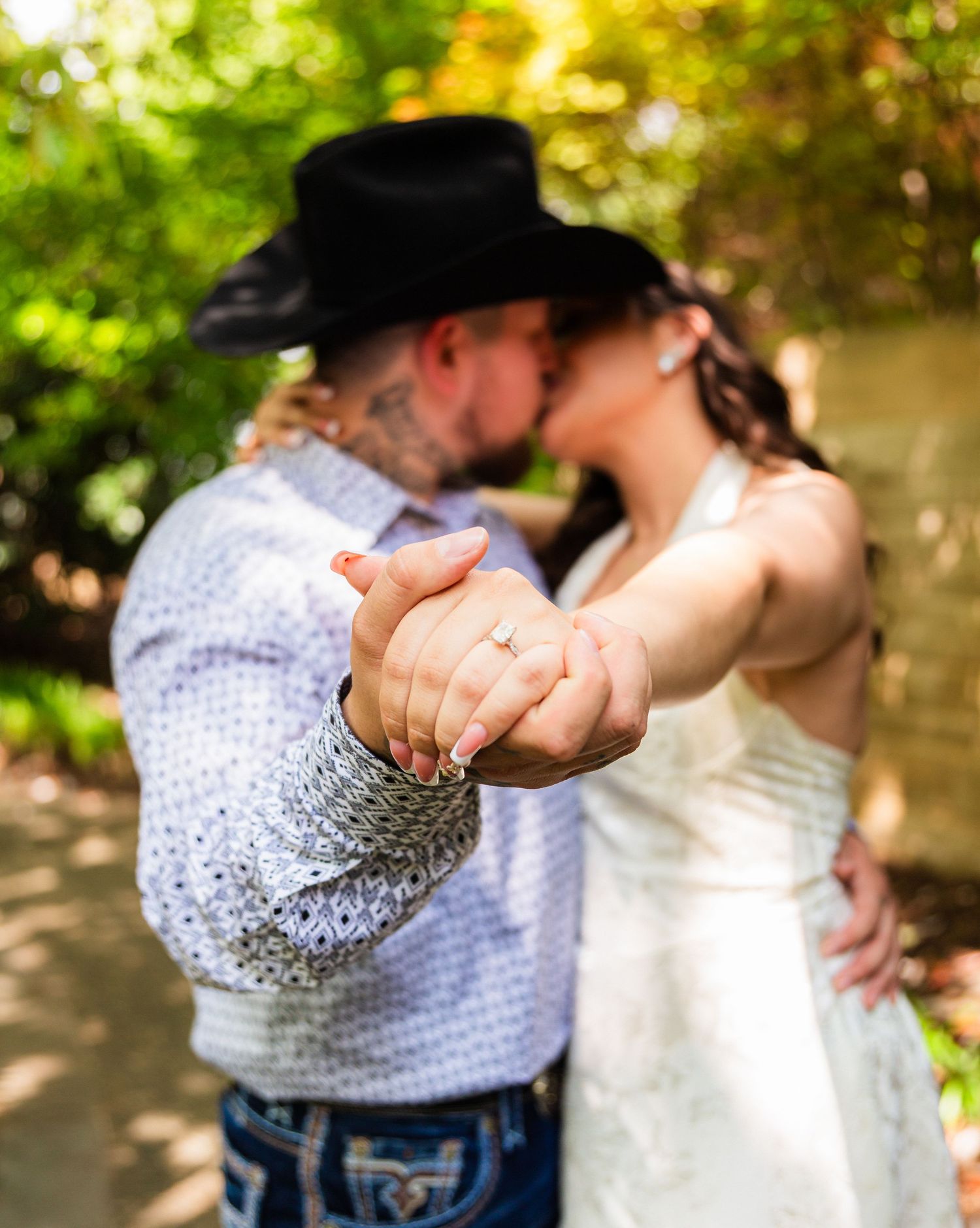 Couple kissing outdoors, man in black cowboy hat and tattoos, woman in white dress, holding hands showing engagement ring.