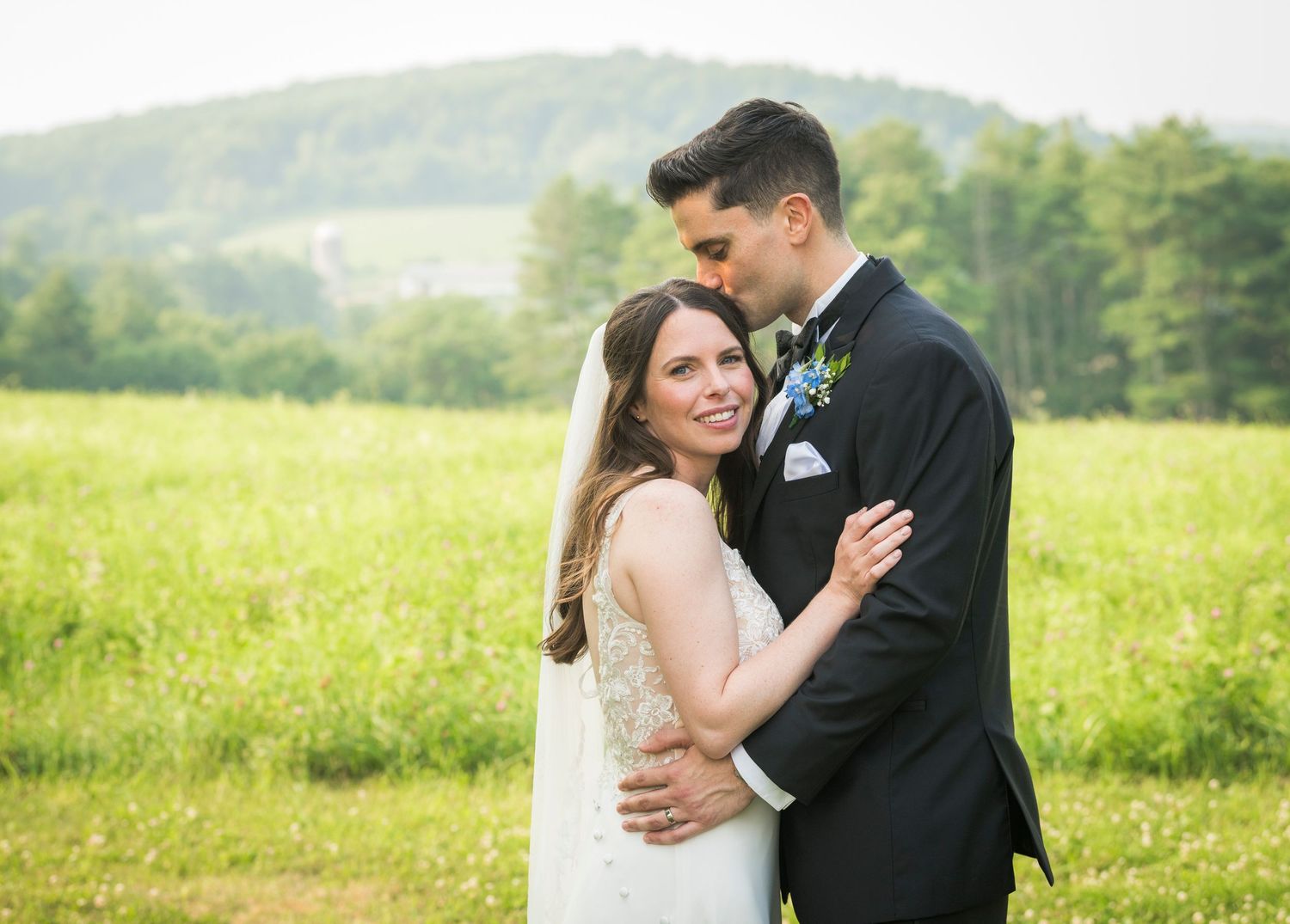 Newlyweds share an intimate moment outdoors with autumn foliage and rustic architecture in background.
