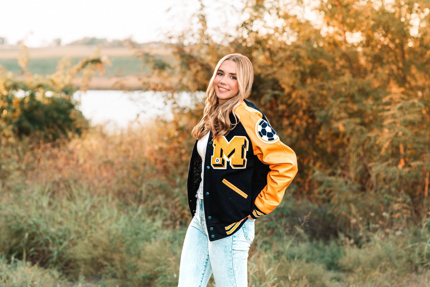 Smiling blonde woman wearing a black and gold varsity jacket with M logo, posing outdoors in golden hour light.