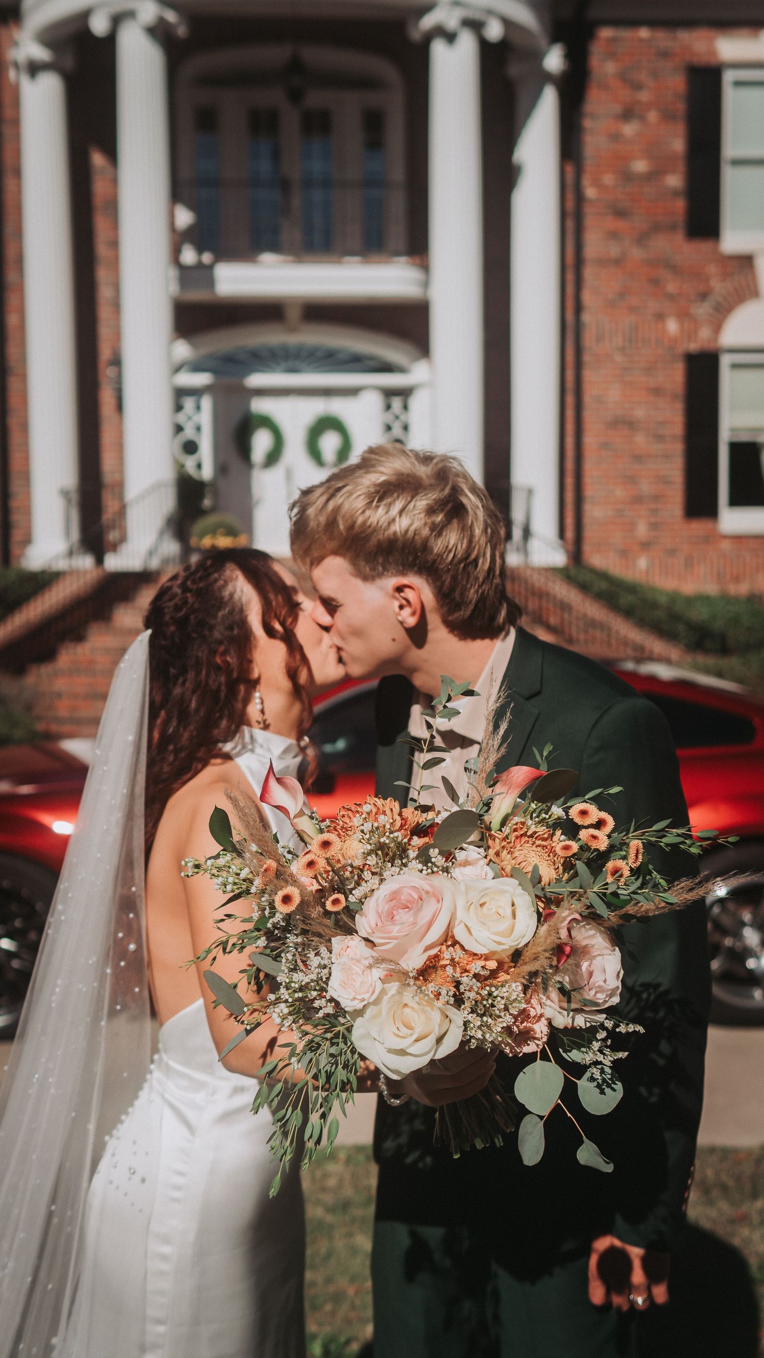 A bride and groom share a kiss while holding a lush floral bouquet outside an elegant brick mansion on their wedding day.