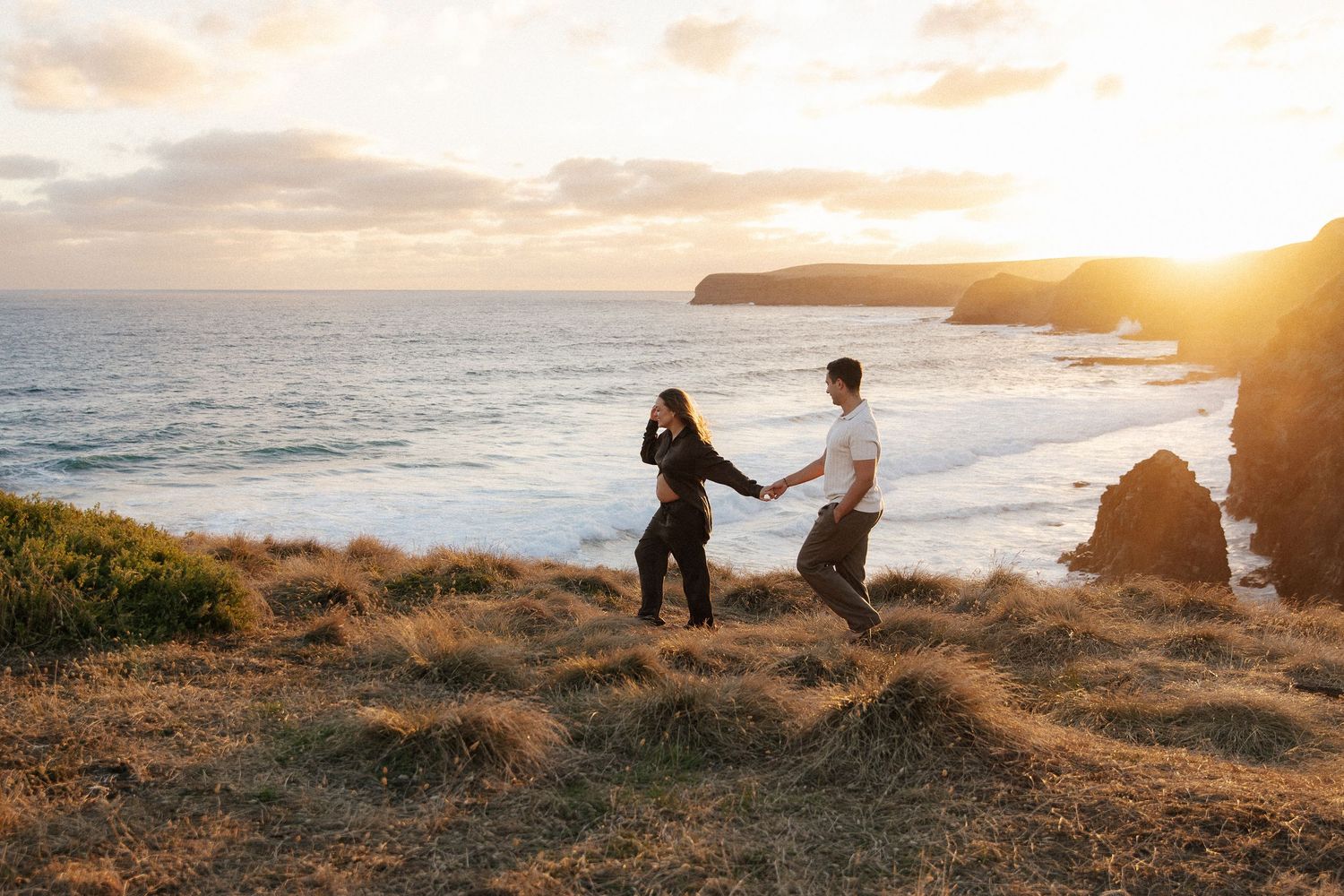 Pregnant woman in black dress smiling at sunset beach, framed by partners hands forming heart shape.