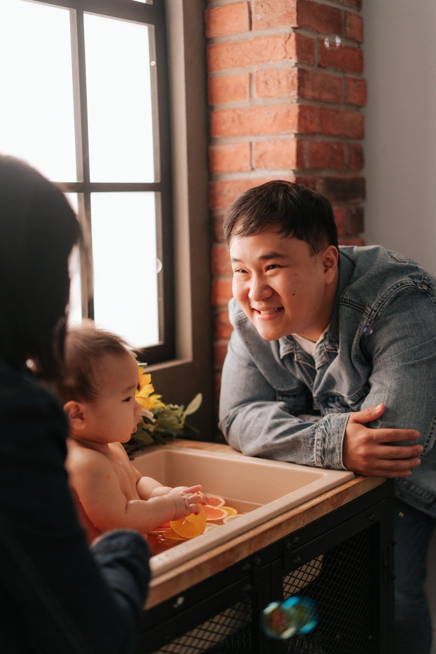Smiling Asian father leaning over baby bathtub while mother tends to infant during bath time near brick wall window.